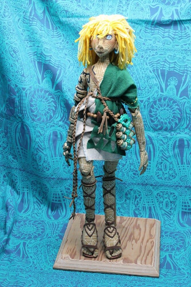 Photo of a mixed-media cloth doll of Link from Legend of Zelda: Tears of the Kingdom. He has yellow wool hair, a painted face, and intricately patterned cloth skin and is wearing a green and white tunic with beaded and braided accessories. 
