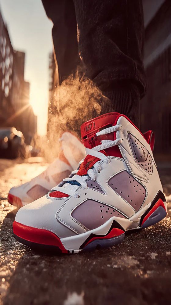 A low-angle shot showcases a pair of Air Jordan 7 Retro sneakers on a wet city street at sunset. The foreground focuses on one sneaker in sharp detail: predominantly white leather with lavender side panels punctuated by perforations. Red accents outline the shoe's sole and collar, while the tongue displays white laces with a red and white diamond pattern and a small red emblem. "Nike" is stitched onto the back of the red ankle strap. The sneaker sits on a reflective gray pavement, catching the warm glow of the setting sun. Another slightly blurred sneaker is visible behind it. Behind the sneakers, a person wearing dark denim or jean-like pants with a ribbed cuff around the ankle stands out of focus. The urban backdrop features tall buildings lining the street, bathed in the golden light of the setting sun which creates bright glares. The overall atmosphere is warm and urban, with a focus on high-fashion footwear.