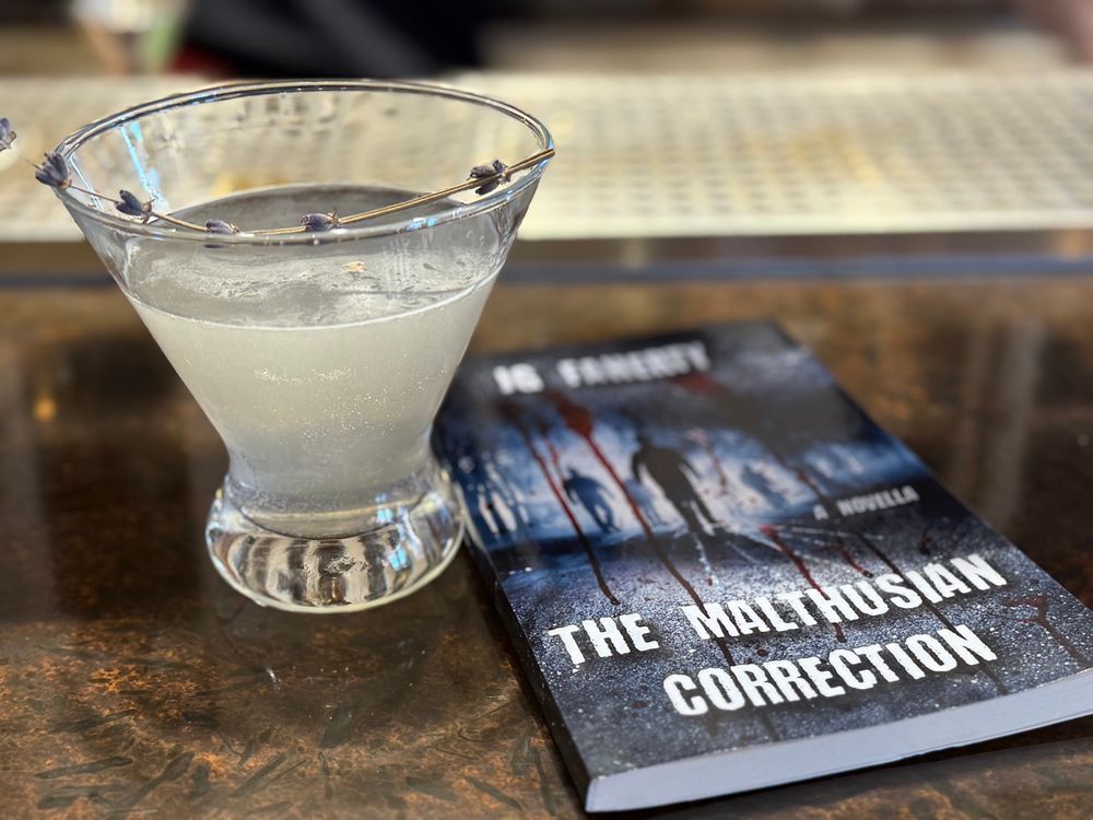 An attractive floral martini perches next to a book on a bar. The book has a dark, creepy cover. It’s The Malthusian Correction by JG Faherty.