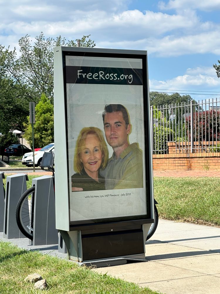 An advertisement for FreeRoss.org next to a rental electric bike stand on a sidewalk along South Dakota Avenue in Washington, DC. A snapshot of Lyn and Ross Ulbrecht appears on a white background. In a black strip above the photo is the website address. 