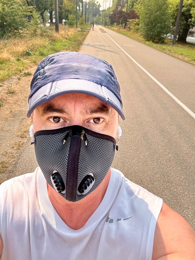 A close-up selfie of a man outdoors on a paved path, wearing a grey ventilated face mask with two filter valves, a light blue cap, and a sleeveless white shirt with visible sweat. The background shows a long straight trail lined with grass, trees, and power poles, with another person walking in the distance under hazy skies.