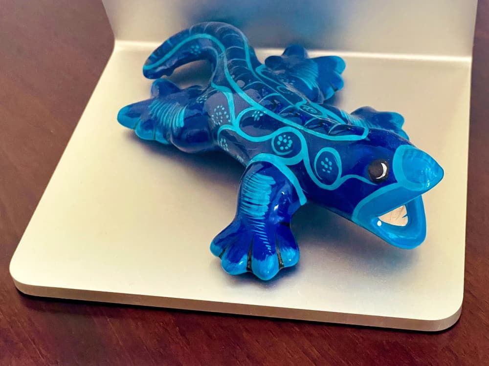 Photo of a blue ceramic figurine in the shape of a salamander, decorated with light blue patterns, which include lines and dots that accentuate its body and limbs. The salamander’s open mouth adds character, making it appear as if it’s mid-motion. The glossy, translucent surface suggests it’s crafted from a smooth, polished material. The salamander is placed on an aluminum platform, the stand of an Apple Studio Display on a dark wood desk, contrasting its vibrant blue against a neutral background.