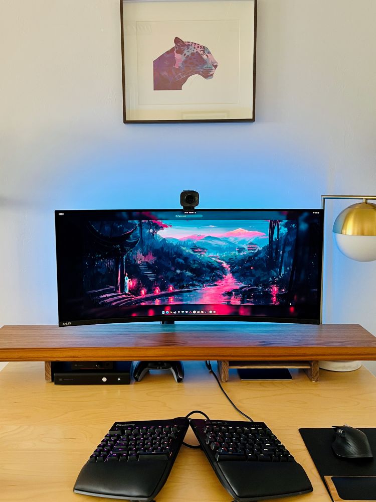 Front view of a wood shelf sitting on top of a desk below a monitor, with audio gear and a game controller tucked neatly inside. 