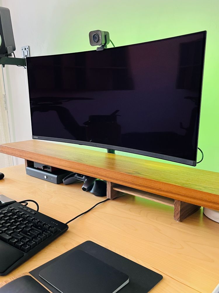 Side view of a wood shelf sitting on top of a desk below a monitor, with audio gear and a game controller tucked neatly inside. 