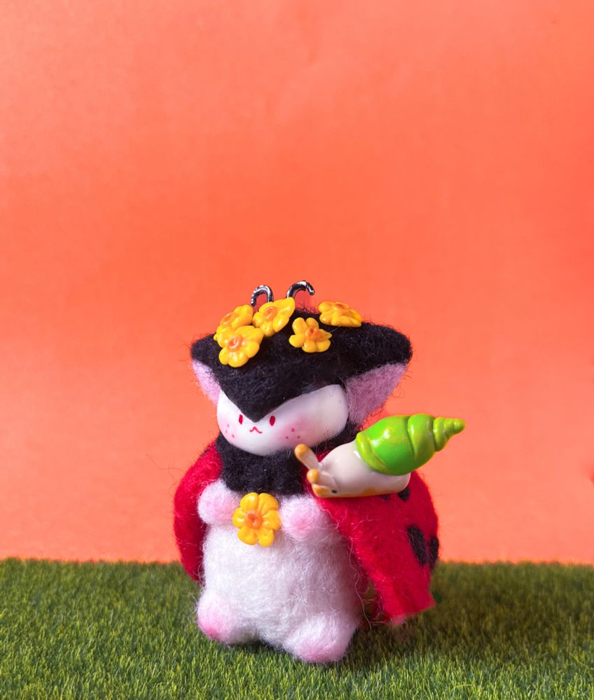 A needle felted cute fantasy creature figure, with a ladybug look, decorated with yellow flowers and a little clay snail on its shoulder.