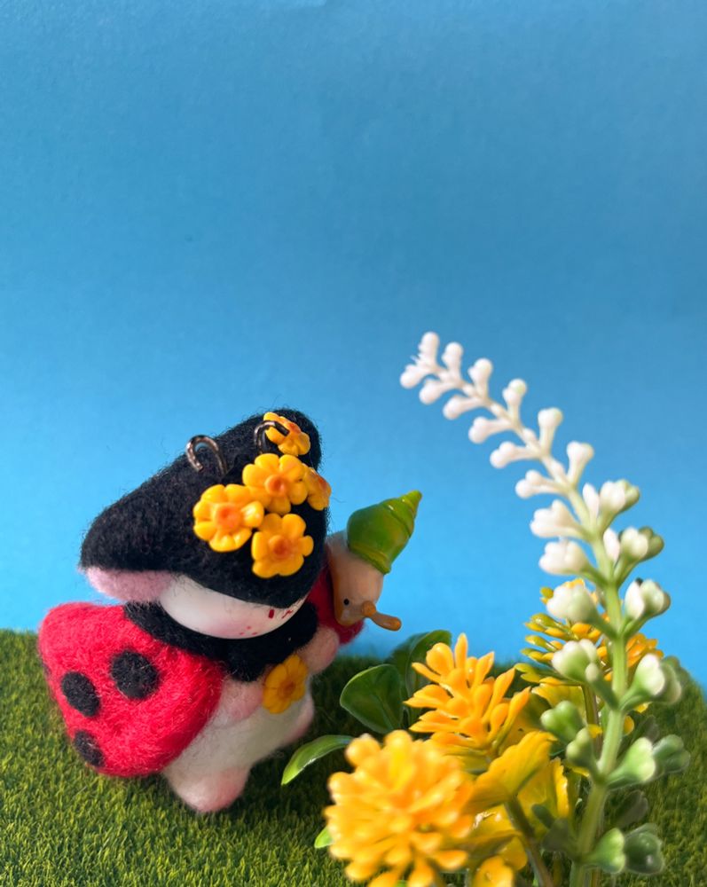 A needle felted cute fantasy creature figure, with a ladybug look, decorated with yellow flowers and a little clay snail on its shoulder.