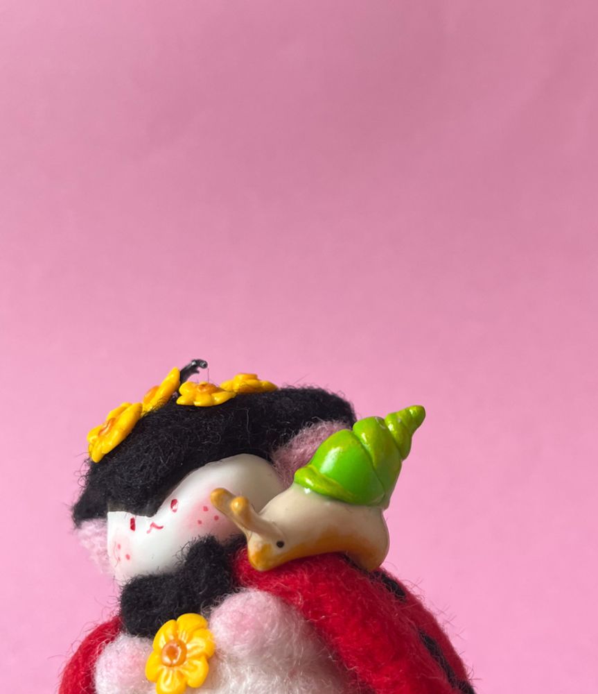A needle felted cute fantasy creature figure, with a ladybug look, decorated with yellow flowers and a little clay snail on its shoulder.