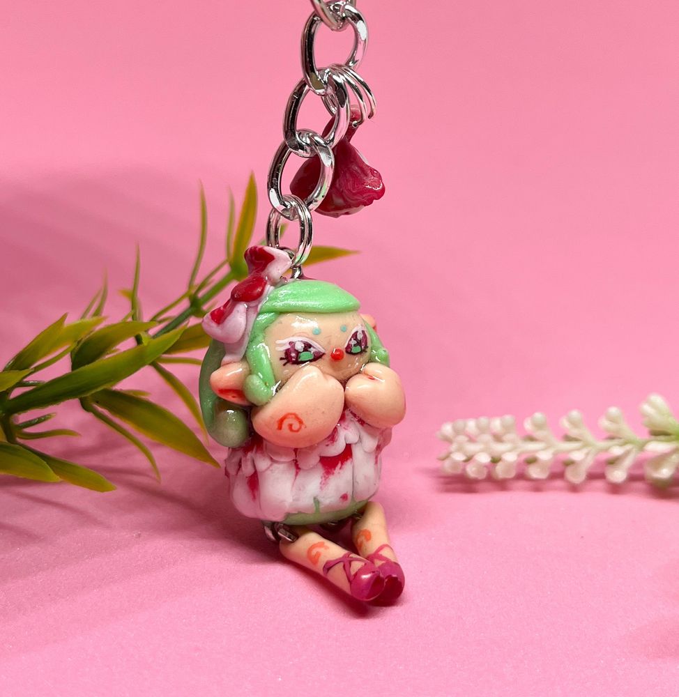 A elf figure holding their hands up to their face sitting on the ground dressed in pink and white with green hair and big eyes in front of a pink backdrop 