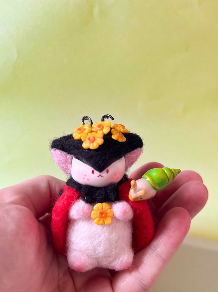 A needle felted cute fantasy oc figure, with a ladybug look, decorated with yellow flowers and a little clay snail on its shoulder.