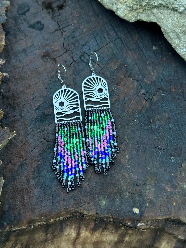 Beaded earrings on a stainless steel frame. Frame has outline of sunset and hills. Bead colors of black, green, pink, purple, blue, and light blue. Bead colors flow together and inspired by aurora borealis.