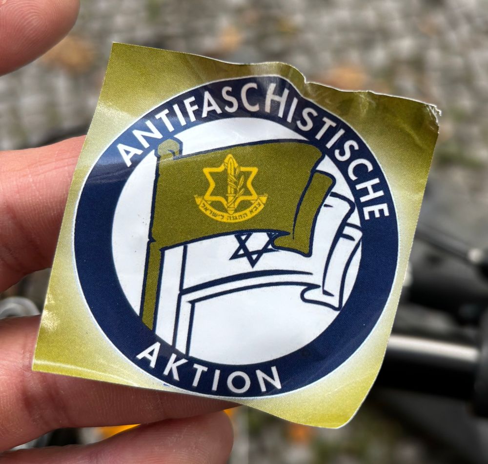 A picture of a sticker found in Berlin, featuring the word “Antifaschistische Aktion” in bold white letters on a dark background. Below it is the emblem of the Israeli Defense Forces (IDF): a yellow Star of David containing a sword wrapped in an olive branch, with Hebrew text at the bottom. The Israeli flag is also visible beneath the emblem, on a white area of the sticker.