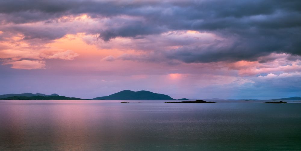 Pink and orange sunset clouds peep through dark blue heavy clouds giving off reflections on a calm sea. Midway in the image are low dark hills and islands scattered horizontally.