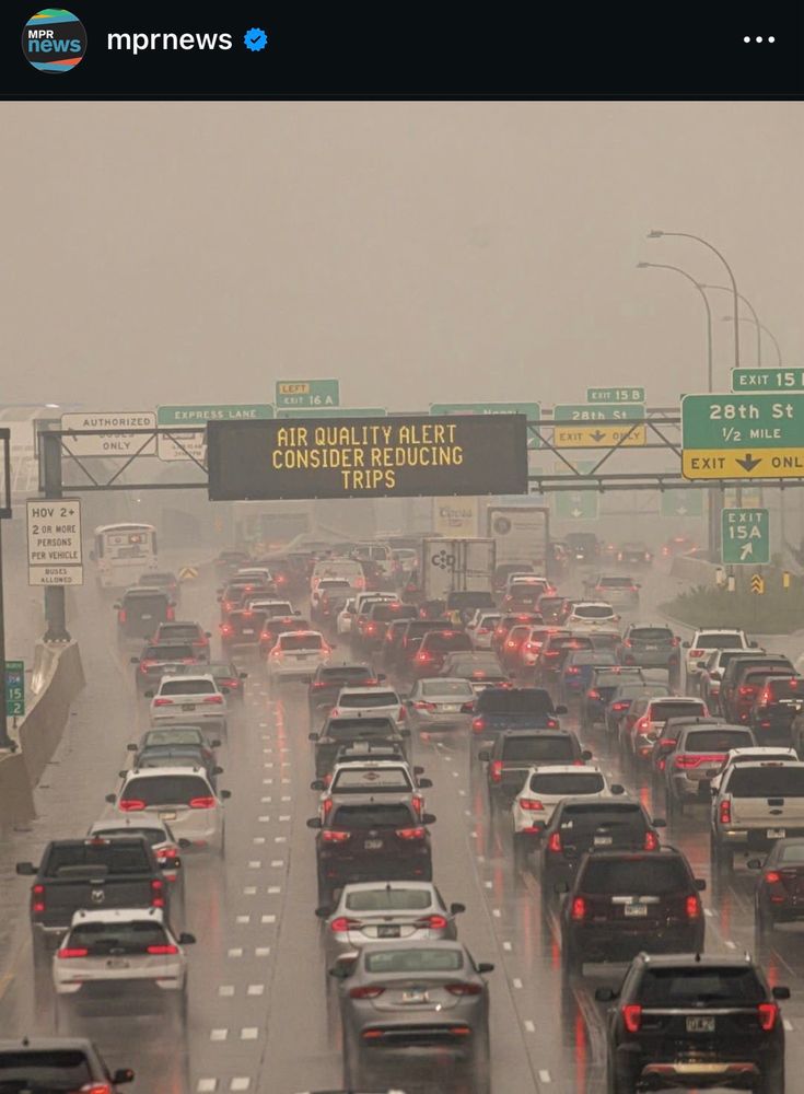 Image shows hundreds of cars slowed/stopped on a highway with signage saying “air quality alert consider reducing trips.” Heavy wildfire smoke is in the air.