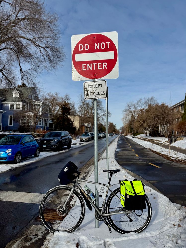 Silver bike parked in front of a sign post. The signs say “Do not enter except bikes” and have clearly been run over by a driver of a car in the past.