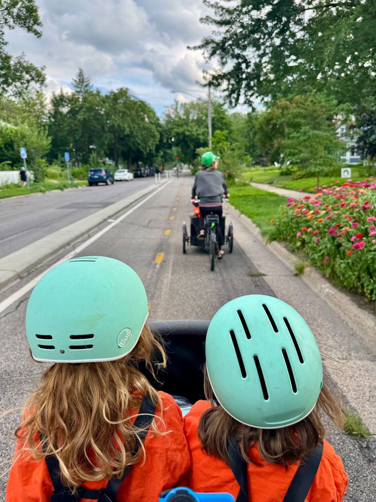 Laura’s kids sit in the front cargo area of their Bunch bike. Another parent is riding a different cargo trike with kids right in front. They’re riding in a 2 way curb protected bikeway.