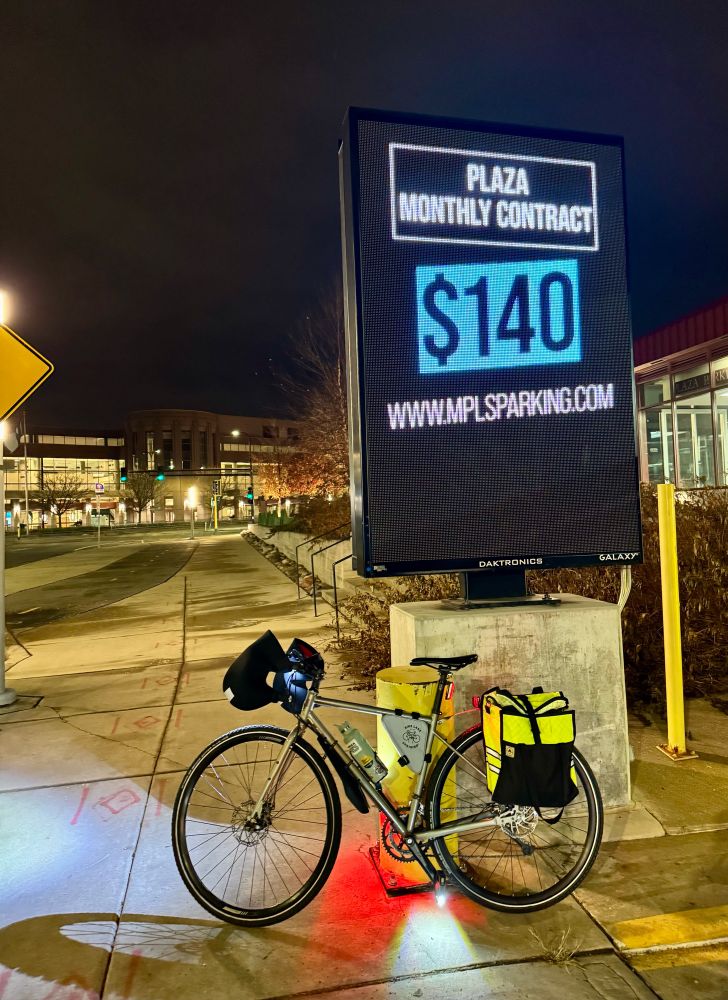 Bike parked next to a protected bikeway, in front of a sign, at night. 

Sign says:
“Plaza monthly contact 
$140
Www.mplsparking.com”