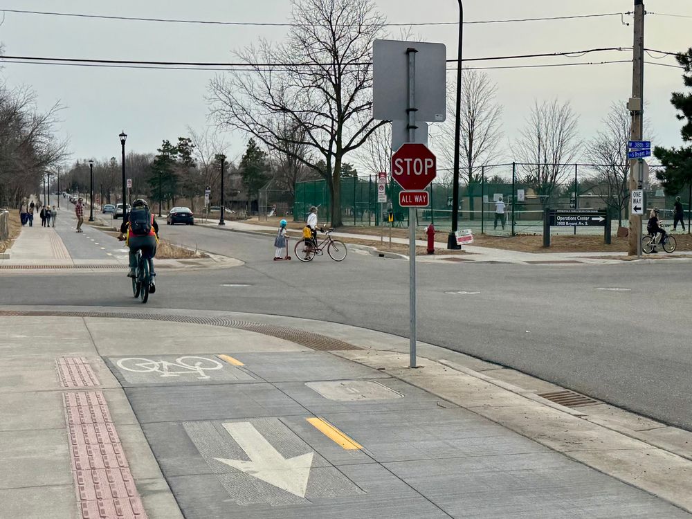 Intersection of a well designed street with a sidewalk level 2 way bikeway, narrow lane for cars, and sidewalks on both sides. Many people are walking, biking, scootering and skateboarding. No one is driving.