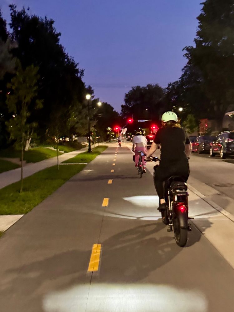 A block of Bryant Ave S at night with 3 people on bikes (+ Laura behind camera) biking, just one car 