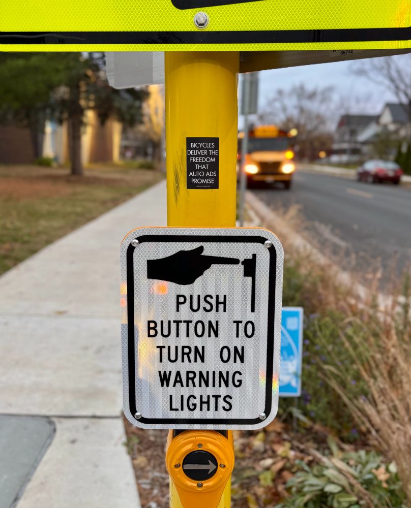 Pedestrian beg button with a sticker on the pole that reads “Bicycles deliver the freedom that auto ads promise”