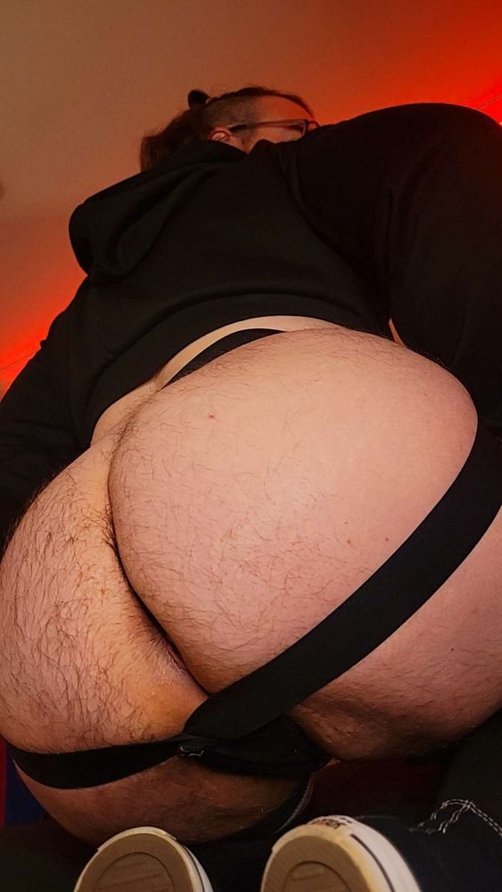 Idiot Angel kneeling on a bed. his round hairy ass in a black jockstrap takes up most of the frame. He leans forward, giving the viewer a peek at his thick thighs. He wearing a cropped black hoodie his long hair in a half up bun. 