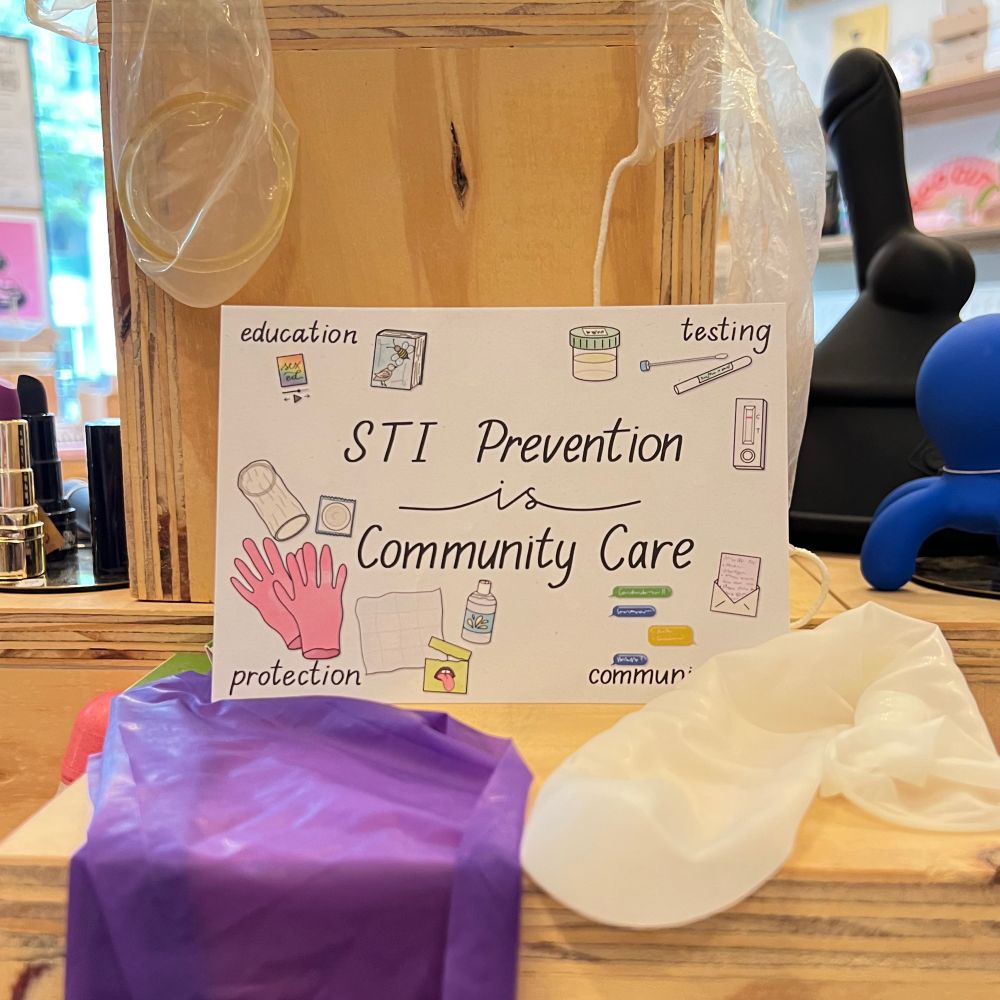 A postcard on a wooden display surrounded by 2 dental dams, an external condom and an internal condom

It has with text in the middle saying: ‘STI prevention is community care’. The 4 corners say:  1) ‘education’ along with a book about birds & bees and an audio interface playing ‘sex ed’ 2) ‘testing’ surrounded by a cotton sample swab, a urine cup and a test card 3) ‘communication’ next to a letter and conversing text bubbles in 3 colors 4) ‘protection’ among gloves, an internal condom, an external condom, a dental dam and a bottle of lube