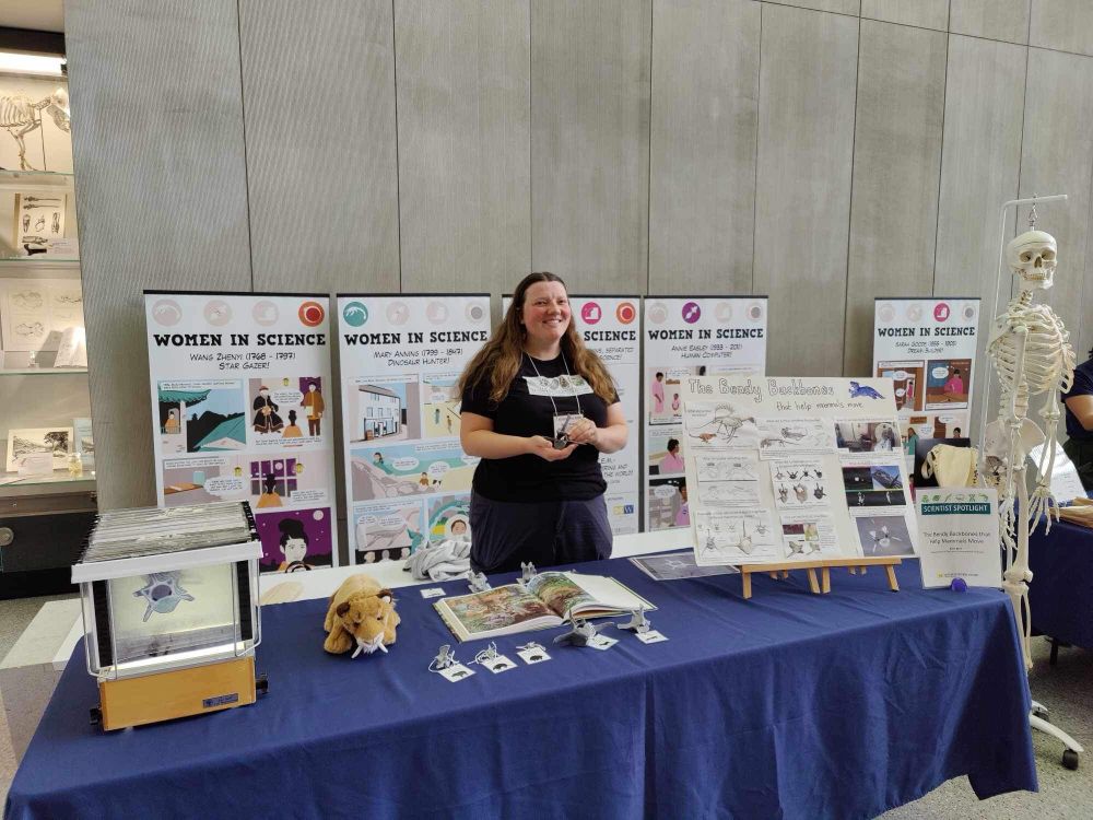 Table with CT scan image, 3D prints of vertebrae, and poster. Woman standing behind the table holding a vertebra 3D print. 