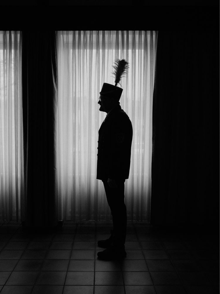 A miner poses sideways for a black and white portrait in front of a curtained window. His silhouette is very characteristic with the bushy shape of the plume on top of his shaft hat.


