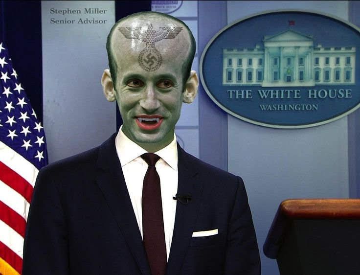 Digitally edited picture of Deputy Chief of Staff Stephen Miller. His skin is vaguely green, he's revealing vampire canines as he smiles, and his forehead is swollen and bears a tattoo of some Nazi falcon swastika garbage icon. He's standing near a podium, a US flag and the seal of The White House.