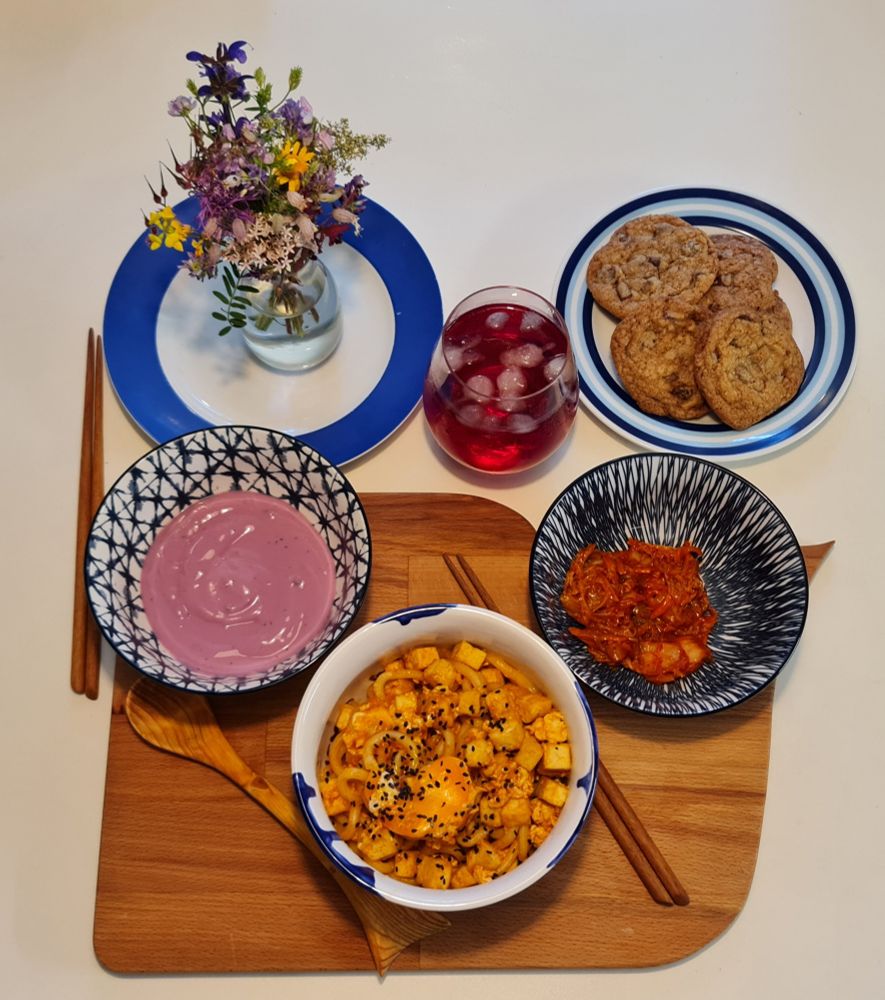 a white table with various bowls of food laid out. some bowls are on a wooden board in the shape of a whale. there is a large bowl with tofu udon stir fry and a smaller blue bowl with some kimchi. another small bowl with purple blueberry yoghurt. behind the board are a glass with a clear, red-ish blackcurrant drink with ice cubes. next to it a plate with espresso chocolate chip cookies. in the left upper corner another plate with a small vase with a colourful wildflower bouquet inside. there are two pairs of wooden chopsticks and a wooden spoon with a handle. the same shape as the whale fin of the large wooden board. 