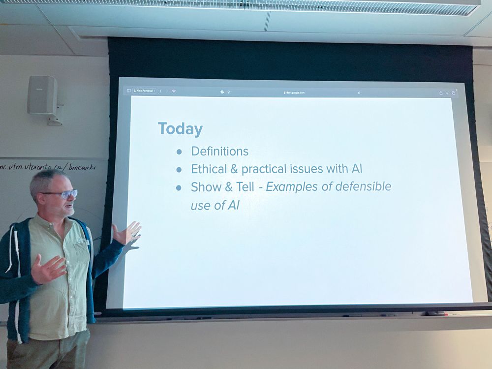 Session co-organizer Nicholas Woolridge who wears glasses and a green shirt with a dark hoodie, gestures in front of a projected slide. The slide, lists topics for discussion, including definitions, ethical and practical issues with AI, and examples of defensible AI use.