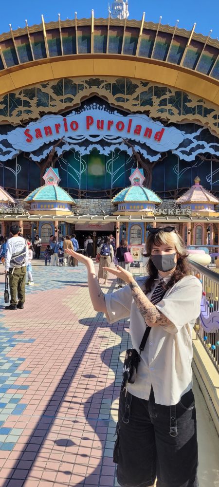a person with a mask stands before the opening of a themepark, sanrio puroland, holding their hands towards the entry