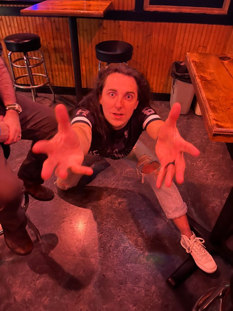A long haired brunette in a cropped black shirt and faded blue jeans in a dive bar on their knees with their hands reaching up to the camera. Their face is alarmed and anguished as if they’re saying “why would you do this to me” 