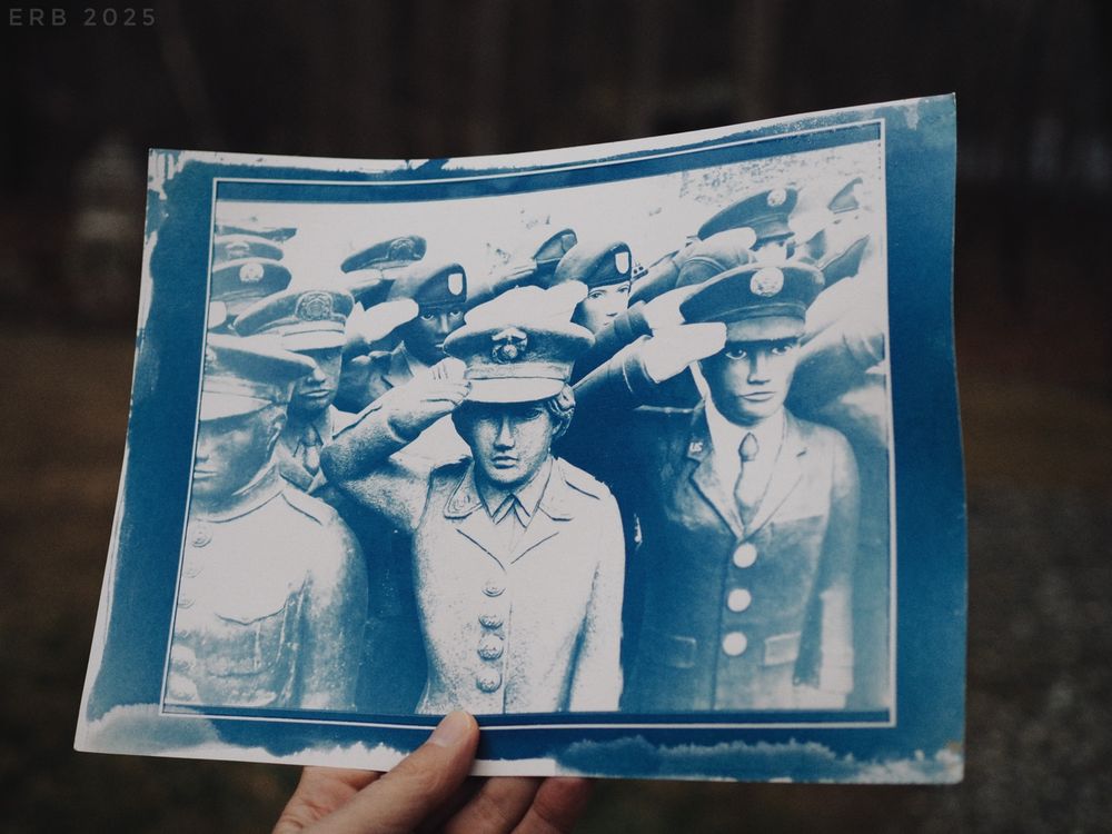 The photograph shows a hand holding a cyanotype print. The print depicts a tight group of soldiers in uniform, all facing forward and saluting. Their faces are serious and stylized, almost like carved figures or mannequins rather than living people. The cyanotype process gives everything a deep blue tone with white highlights, creating high contrast and a slightly ghostly look.

The central figure is a soldier saluting with his right hand, his expression stern. Two other soldiers flank him at close distance, also saluting. Behind them is a dense cluster of additional uniformed figures, their hats and raised hands overlapping so closely that their bodies merge into a pattern of shapes and shadows.

The cyanotype print has irregular dark edges, brush marks, and a border around the image, emphasizing its handmade quality. The background of the larger scene is an outdoor setting, out of focus, with bare trees and muted light.

The overall impression is of a repeated or massed image of military figures, unified in gesture and tone, rendered in a historic photographic process that adds an aged and somewhat haunting quality.