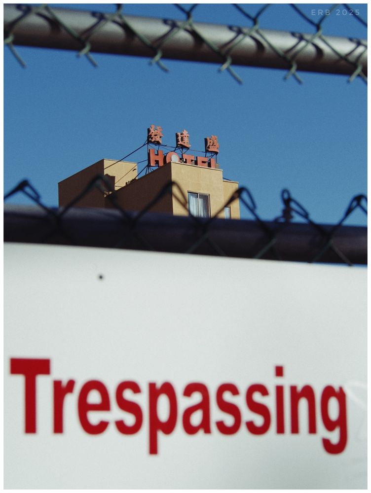 A hotel sign with Chinese characters rises against a clear blue sky, seen through the blurred links of a chain-link fence. A large white sign in the foreground shows the word Trespassing in bold red letters. The frame splits the scene into layers: warning, barrier, and the distant building. The result feels controlled and observational, with a tension between access and exclusion.