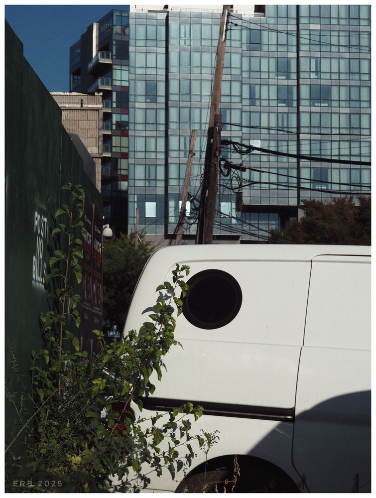 The photograph shows an urban street scene with layers of detail. In the foreground, there is the back portion of a plain white van. A circular black window on the van stands out against its smooth white surface. Next to the van, a leafy green plant grows upward along a dark green wall. On this wall, white stenciled letters read “POST NO BILLS.”

Behind this, wooden utility poles lean slightly, carrying a tangle of overhead wires that crisscross the scene. In the background, a modern high-rise building with a grid of blue glass windows dominates the view, its reflective panels catching the light.

The composition contrasts the rough textures of plants, poles, and wires with the sleek geometry of the glass building.