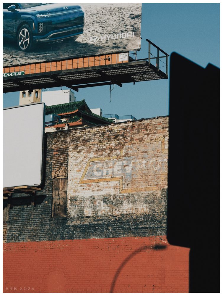 This image shows a layered urban scene made up of geometric shapes, shadows, and textures.

In the upper part of the frame, a modern billboard features a blue Hyundai SUV against a sandy background. Beneath it, the roofline of a building with green pagoda-style tiles peeks out, adding a contrasting architectural shape.

The middle of the image is dominated by a weathered brick wall, where a faded painted advertisement with the word “Chevrolet” and a lightning-bolt motif can just be made out. Time and weather have softened it into a ghost sign, blending with the rough surface of the bricks.

In front, a large dark shape — possibly the shadow of another sign — cuts diagonally into the right side of the frame. At the bottom, a red-painted wall provides a solid block of color, interrupted by the curved shadow of a streetlamp.

The photograph flattens these elements into an abstract composition of rectangles, diagonals, and curves, where the subject matter is secondary to the play of lines, tones, and surfaces.