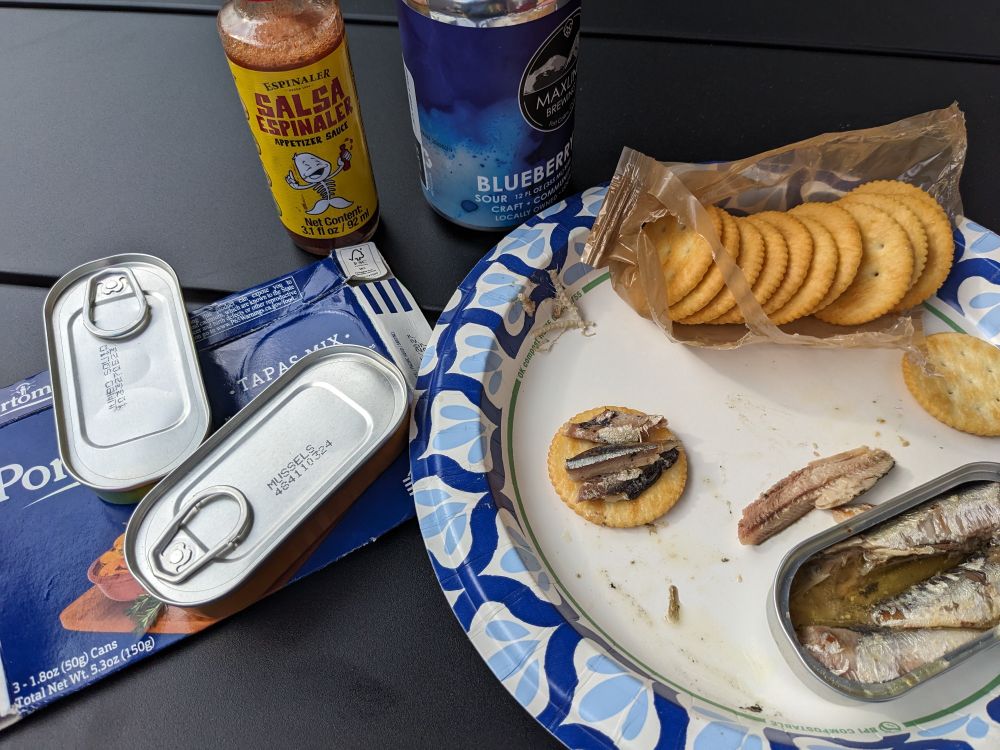 Box of tapas mix, which includes small sardines, squid in garlic and mussels in flavored pickled sauce. Bottle of Espinaler hot sauce. Can of blueberry lime sour beer. Plate with crackers and an open can of sardines.