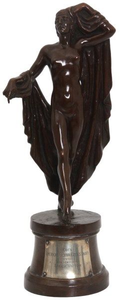 A bronze female nude holding a cloth draped over her right hand/forearm and raised left arm, with a brown patina, stands on a brown award platform. The silver plaque on the platform says the award was presented to Sally Victor in 1956.
