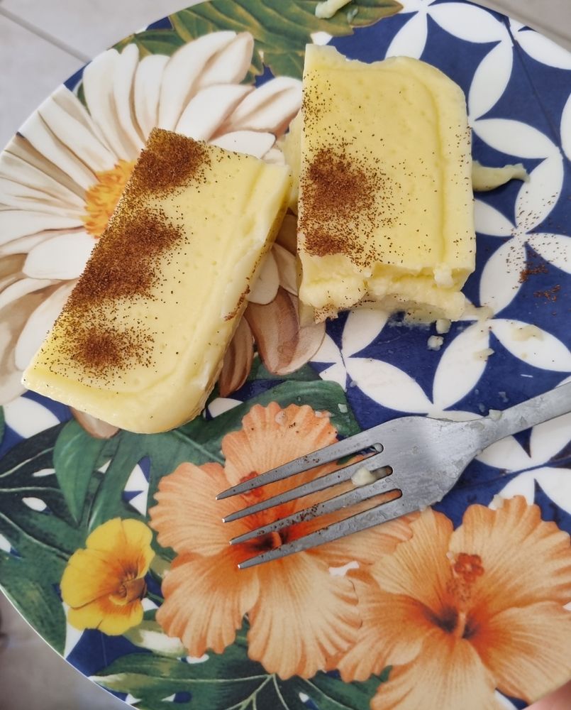 dois pedaços de canjica/mingau de milho num prato de sobremesa estampado com azulejos azuis e brancos e flores de hibisco/graxa em tom pêssego, e um garfo

a canjica é amarelinha clara, salpicada com canela