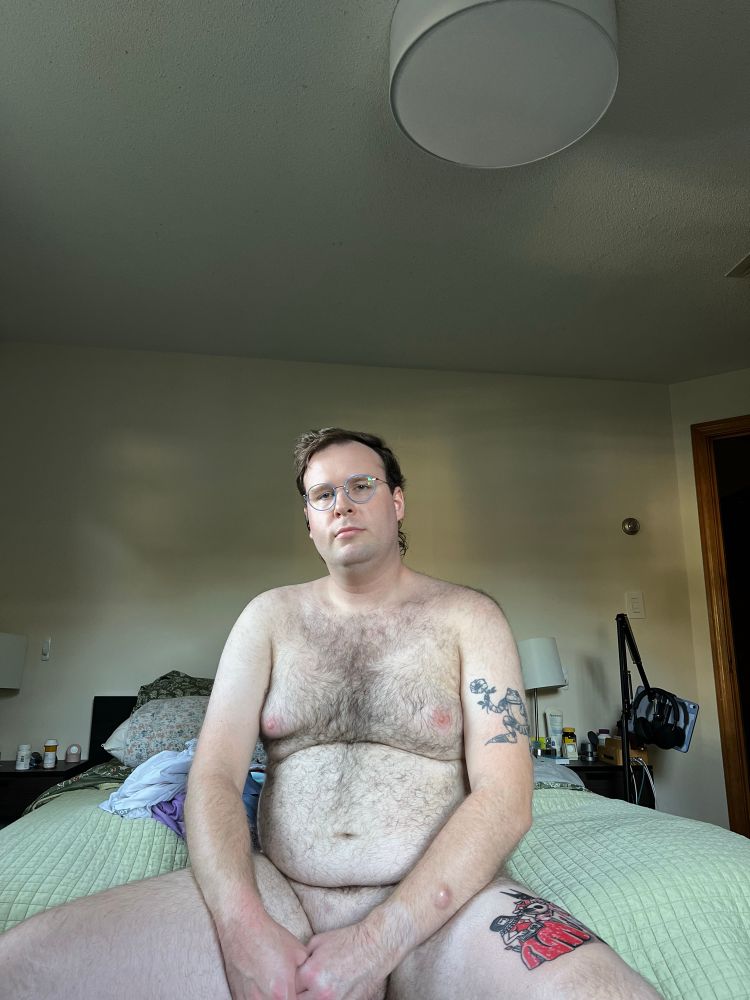 A white masc person sits posed on a bed, naked 