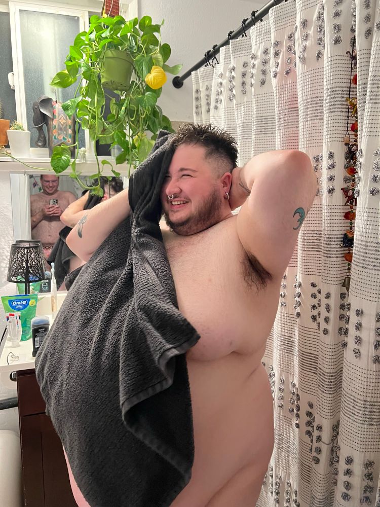A fat masculine person drying off after a shower, and a smaller masculine person in the mirror behind them 