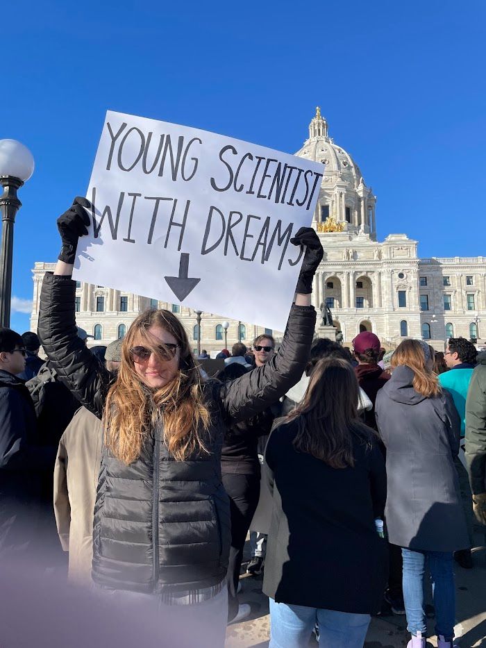 woman holding up a sign that says "Young Scientist with Dreams" in front of a crowd at the Minnesota State Capitol