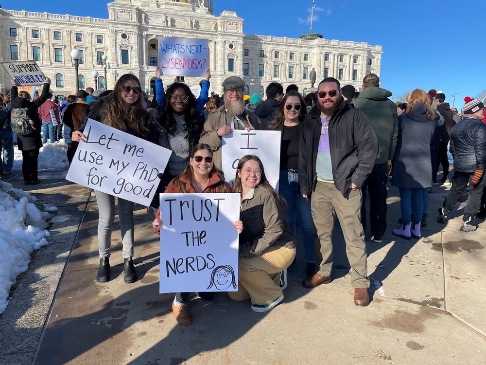 A group of scientists with signs at the Minnesota State Capitol. The signs read "Let me use my PhD for good" and "Trust the Nerds."
