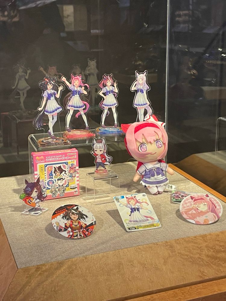 Various pieces of uma musume merch in a display case. There is a haru uraru plushie, a haru uraru pin, a haru uraru trading card, standees of various characters, though the writer of this text only recognizes haru urara, gold ship, and Rice Shower. There is also a Kitasan Black pin. 