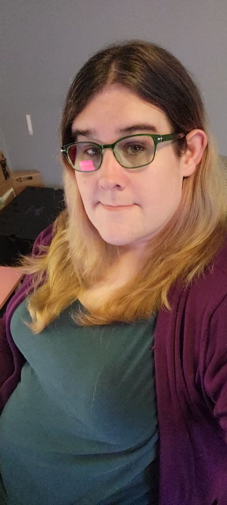 A woman in a teal shirt and purple cardigan, with long hair that was previously dyed leaving a bleached ombre look, sitting at her desk.