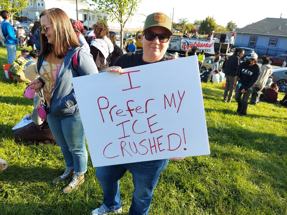 A protester in a crowded park with a sign that says "I prefer my ICE crushed!"