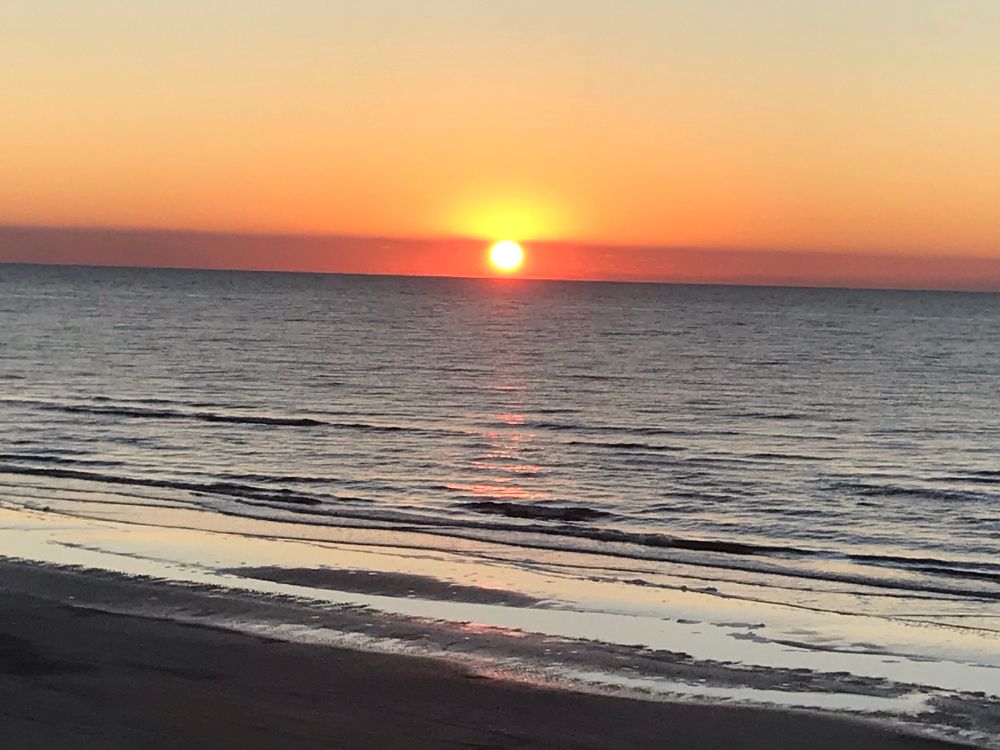 Sunset on the Gulf of Mexico, near Galveston, Texas. A line of thin purple-brown cloud adds a golden glow to the top of the sun just touching the horizon, as the blue waves spread to silver on the dark sand of the shore.
