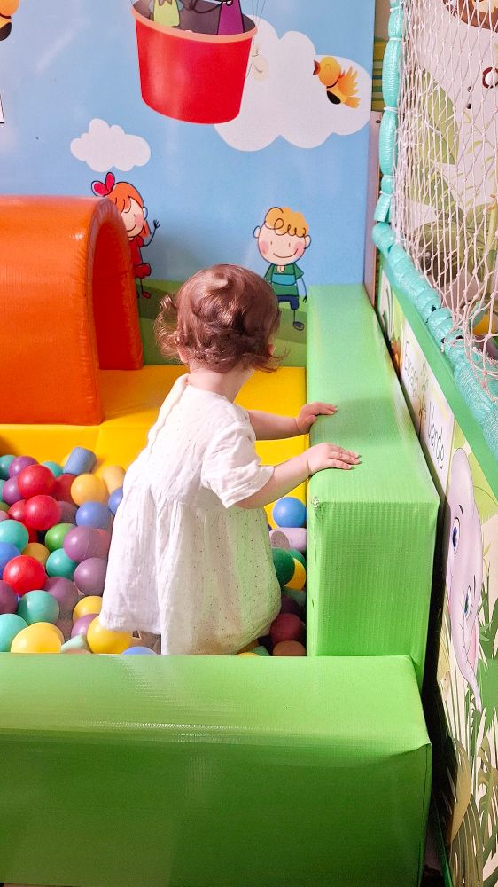 Neném de um ano e meio, de vestido branco e cachinhos caramelo, dentro de uma piscina de bolinhas, apoiando-se na borda com suas mãozinhas. Ela está de costas e não é possível ver seu rosto.