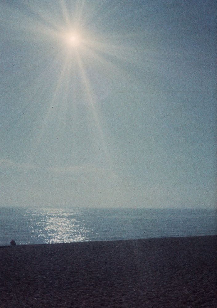 The sun is at about 11 o’clock in this picture in a clear blue sky, which takes up the top half of the pic. Rays radiate from it and light reflects in a vertical stripe from the band of darker blue which comes next, which is the sea. The reddish band below that is the shingle beach.
