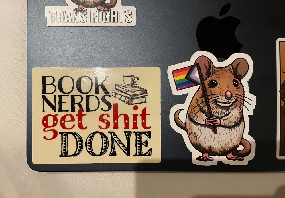 Partial image of the back of a laptop showing various stickers, including one that reads "book nerds get shit done" and one of a cartoon mouse on its hind legs holding a progress pride flag.